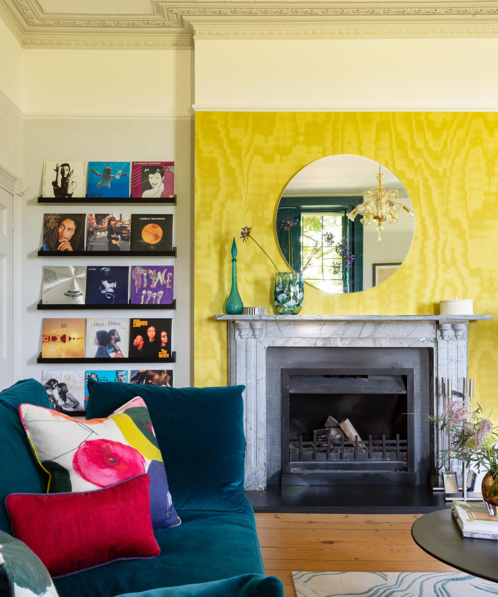 A living room with a bright yellow feature wall behind a marble fireplace. A round mirror reflects light above the mantel. A deep teal sofa with colourful cushions sits nearby, with vinyl records displayed neatly on wall shelves to view.