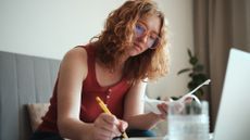 Young woman wearing glasses calculating student loans, making notes in front of her laptop
