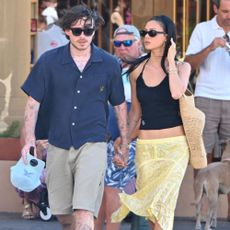 Nicola Peltz Beckham walks down the street wearing a black tank top with back wedge flip-flops and a yellow skirt.