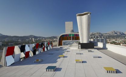 Square-shaped structures are set on the rooftop. Each structure has black & white lines on its side, and the top is in different colors - yellow, blue, and white. The structures are lined up to the reflective surface which gives us the impression of infinite rows of blocks.