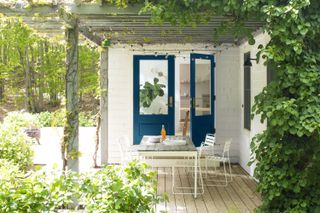 a decked area with a pergola with vines and fairy lights intertwined into it, and planting all around, on the decking and growing up the pergola – with blue doors into the home