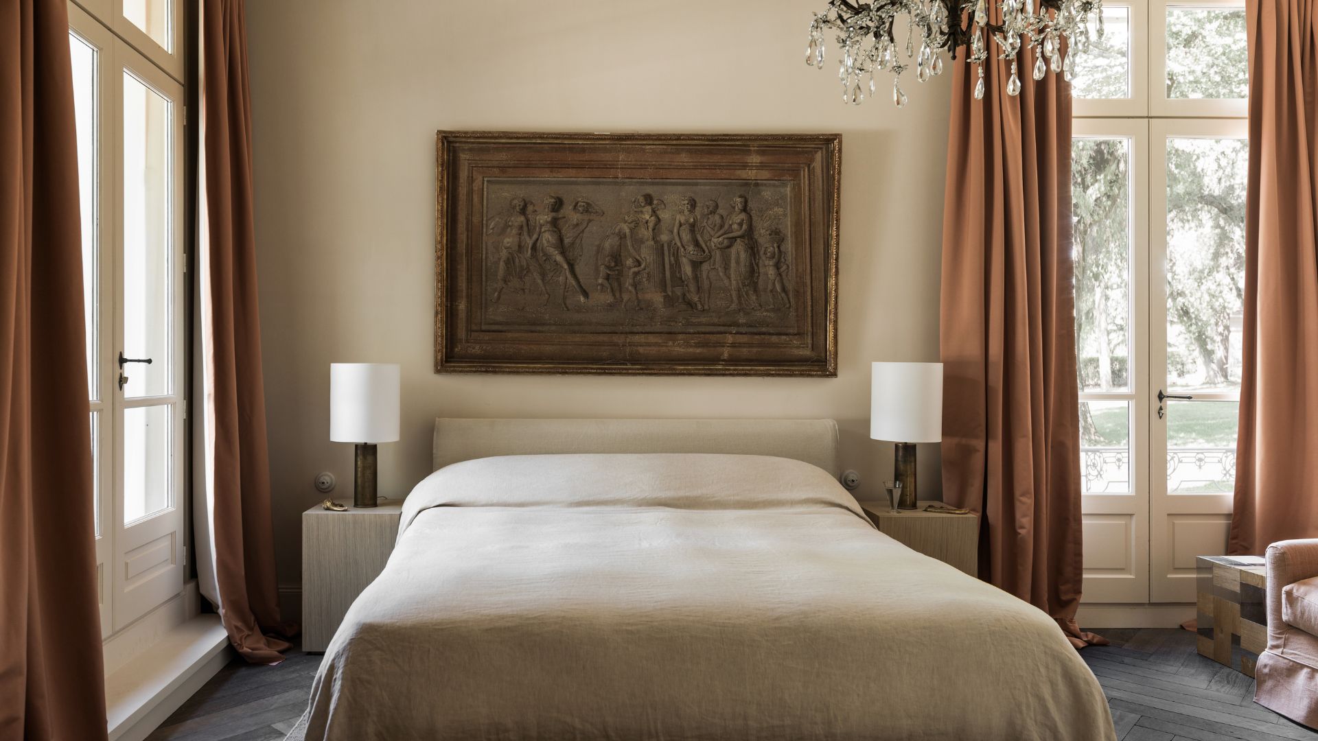 A bright bedroom with a minimalist bed, framed art above the bed, large windowed doors, a chandelier above, and matching nightstands with table lamps