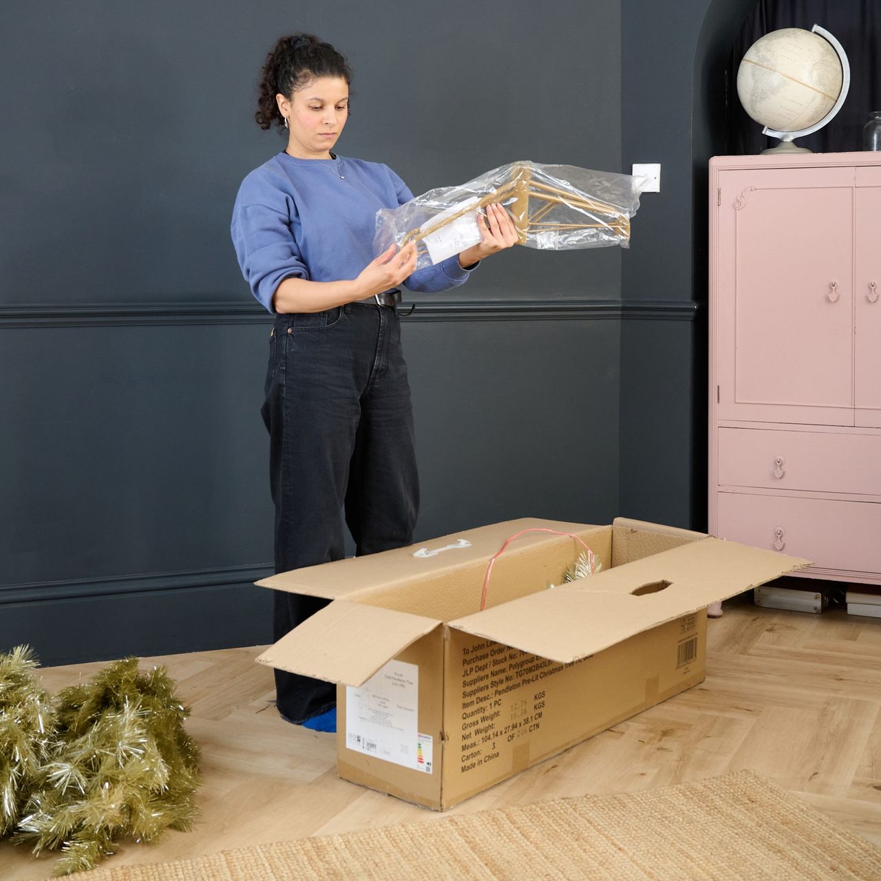 Sara taking out a John Lewis gold Christmas tree and its stand from its box in a black-painted living room