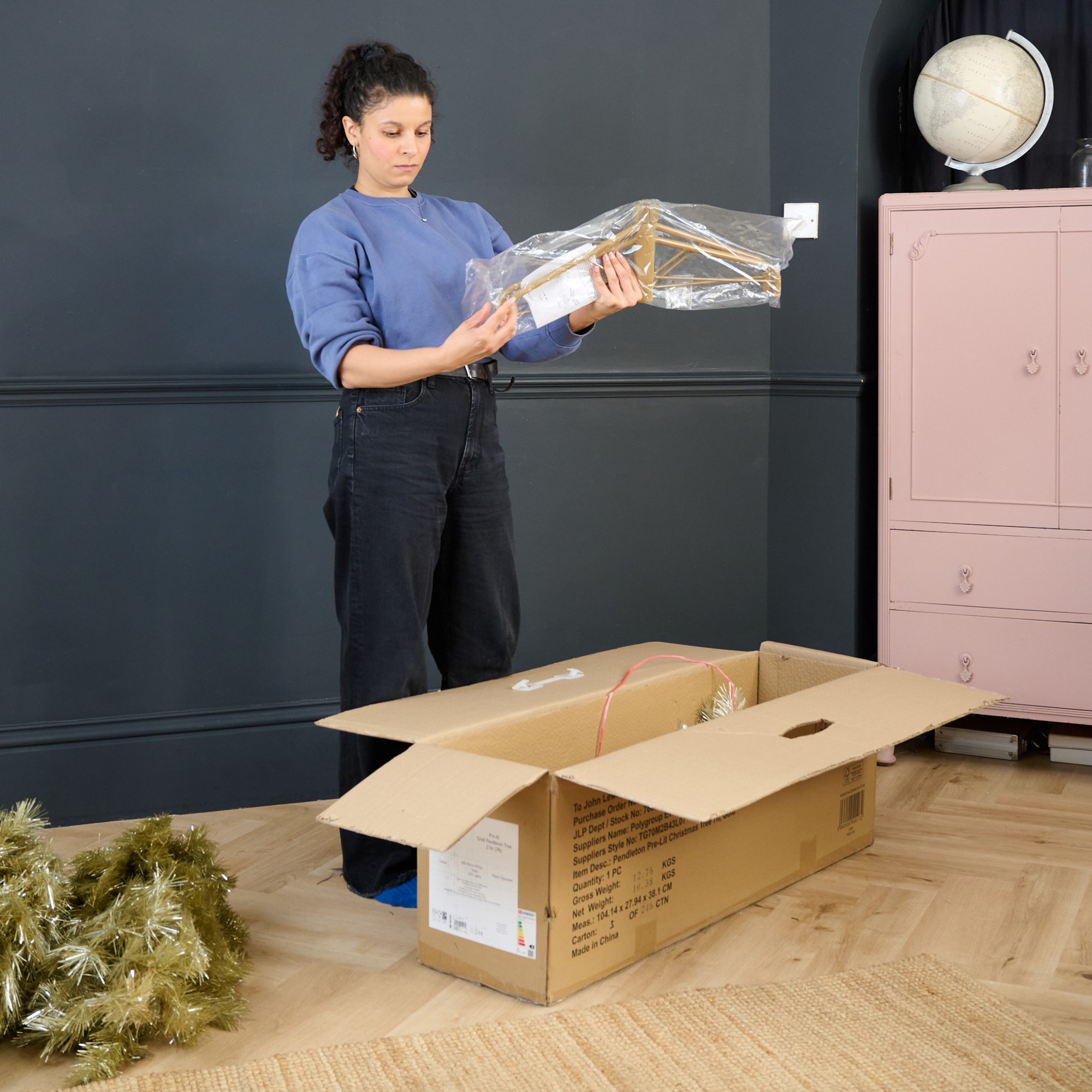Sara taking out a John Lewis gold Christmas tree and its stand from its box in a black-painted living room