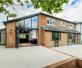 A large semi-detached, brick-clad house which has been extended to the side and rear. The side extension features a sloping roof which has created a large living room with vaulted ceiling inside. There is a large patio running the length of the house.