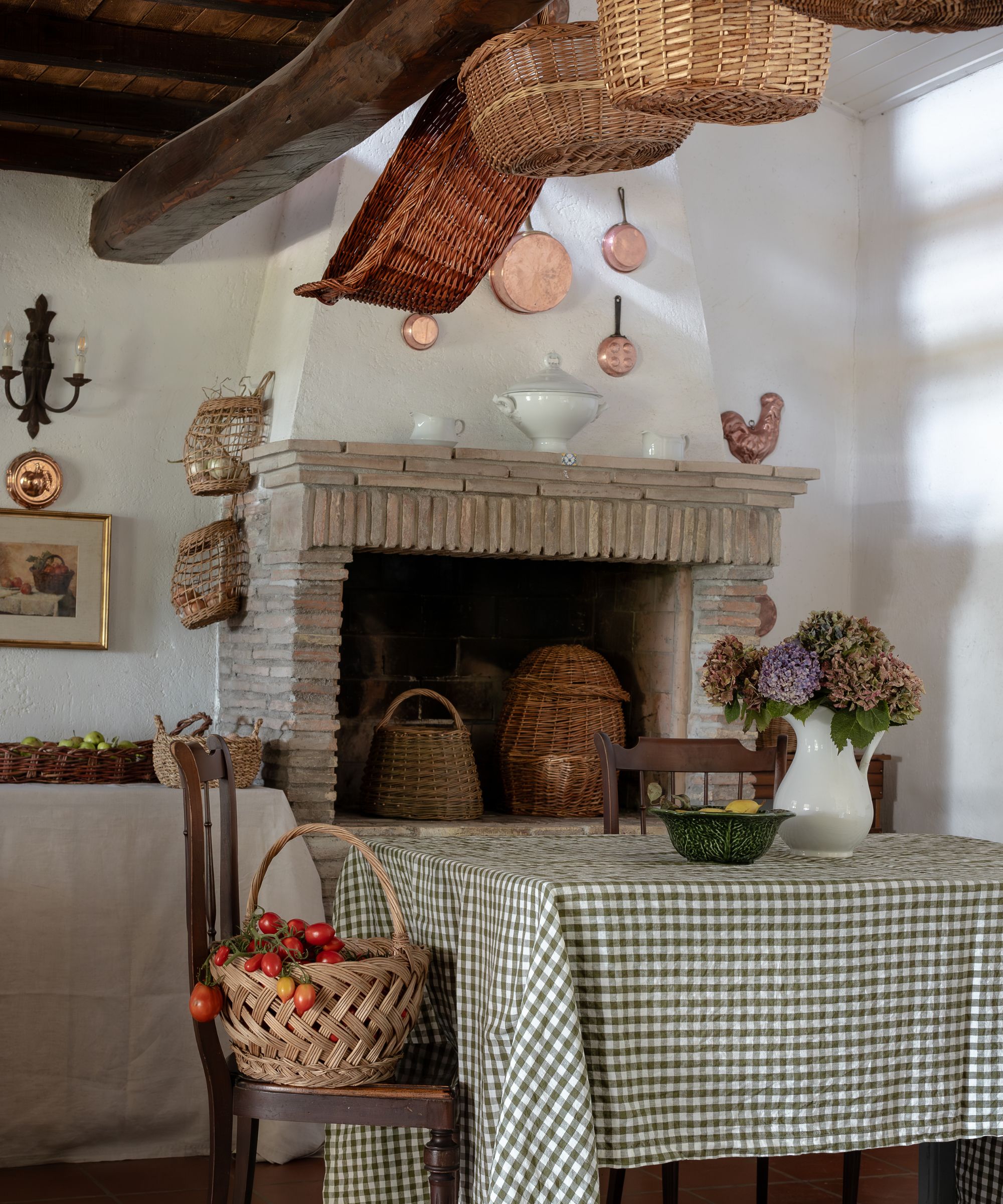 Farmhouse style kitchen with wood beams, large fireplace, table with gingham tablecloth and baskets hanging on ceiling