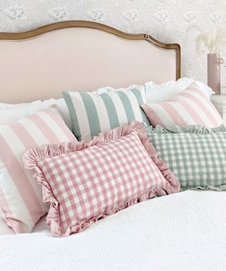 A light pink headboard with a dark wooden trim with white sheets on it and pink and green gingham and striped throw pillows on it