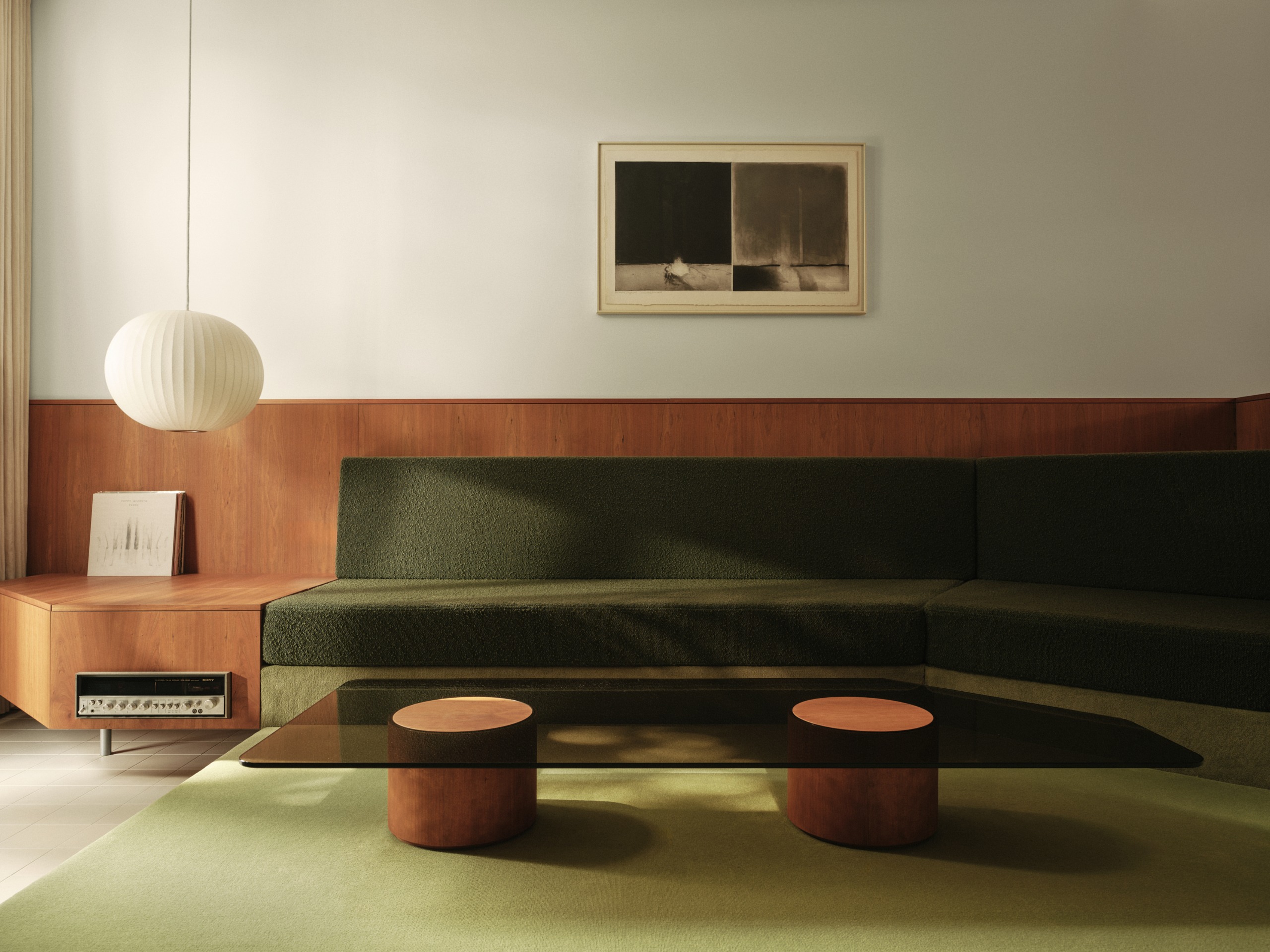 Image of a modern living room with a mid-century style. There is a dark green built-in sofa with a glass and wood coffee table and a pendant light.