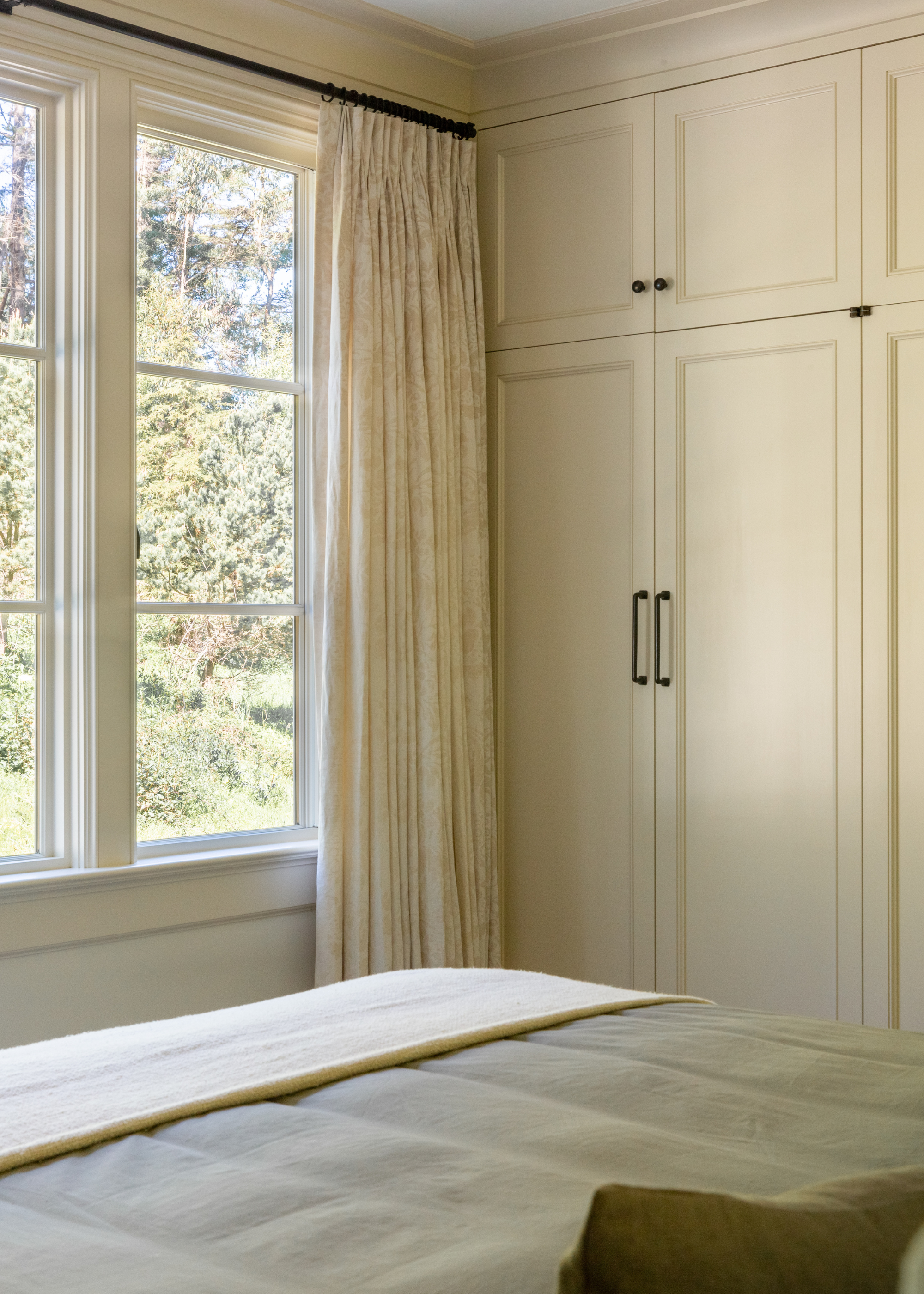 A bedroom with large windows, pleated curtains, a white wardrobe, and a green bedscape