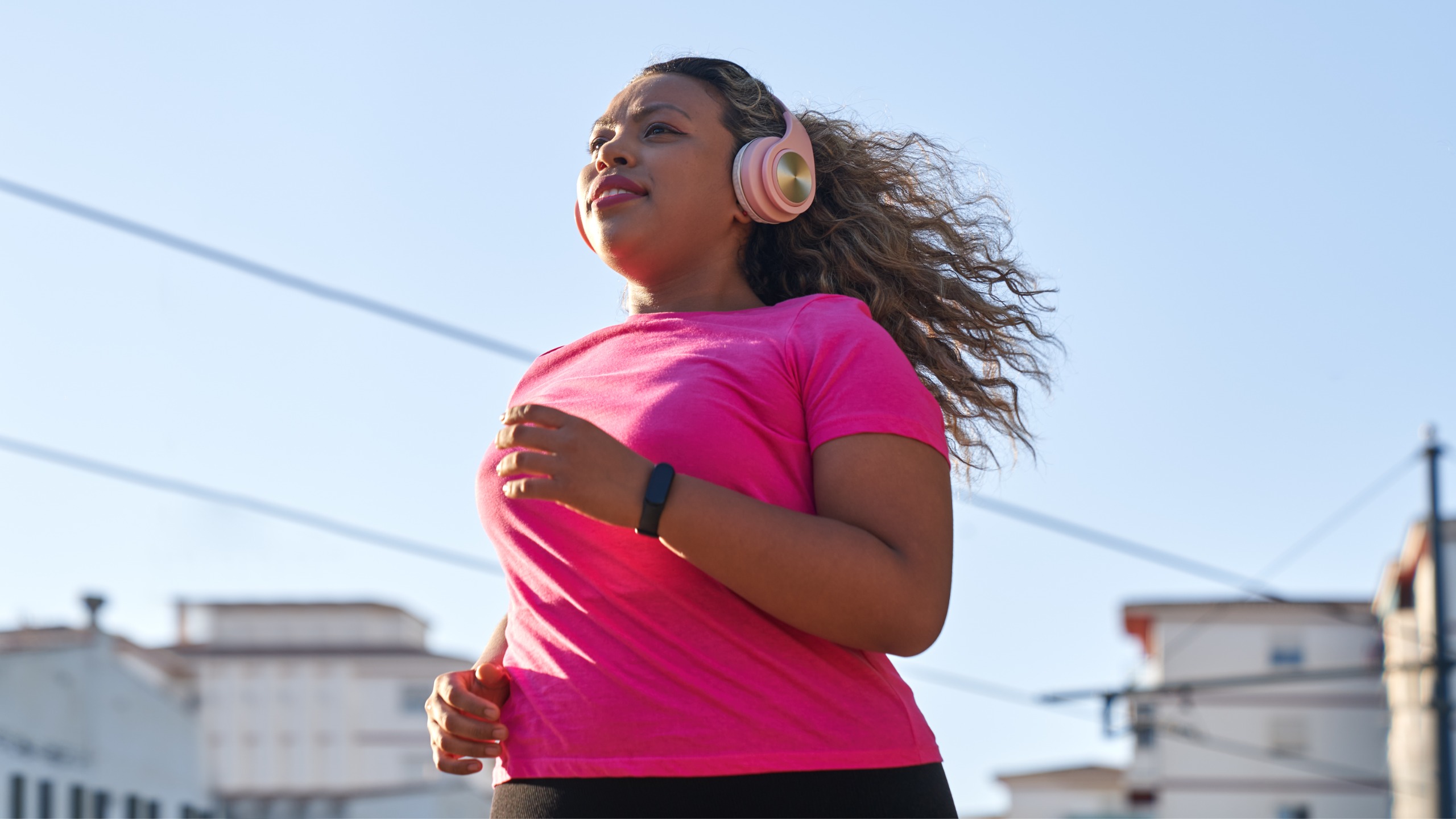 A woman wearing headphones runs along a street. In the background we see buildings and a blue sky.