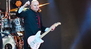 Billy Corgan salutes the crowd. He wears a long black tunic with red buttons and is playing his pink signature guitar from Reverend.