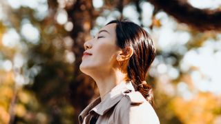 Portrait of young Asian woman having a walk in the park, enjoying the warmth of sunlight on a beautiful Autumn day outdoors and breathing fresh air with eyes closed. Relaxing in the nature under maple trees - stock photo.