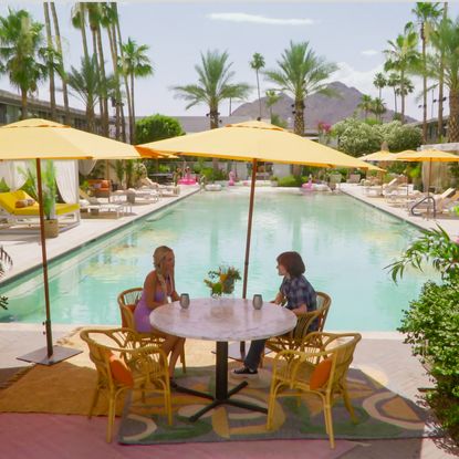 Two people sit at an outdoor table in front of the pool at a motel, with multiple palm trees and a desert mountain in the distance, in 'Sneaky Links.'