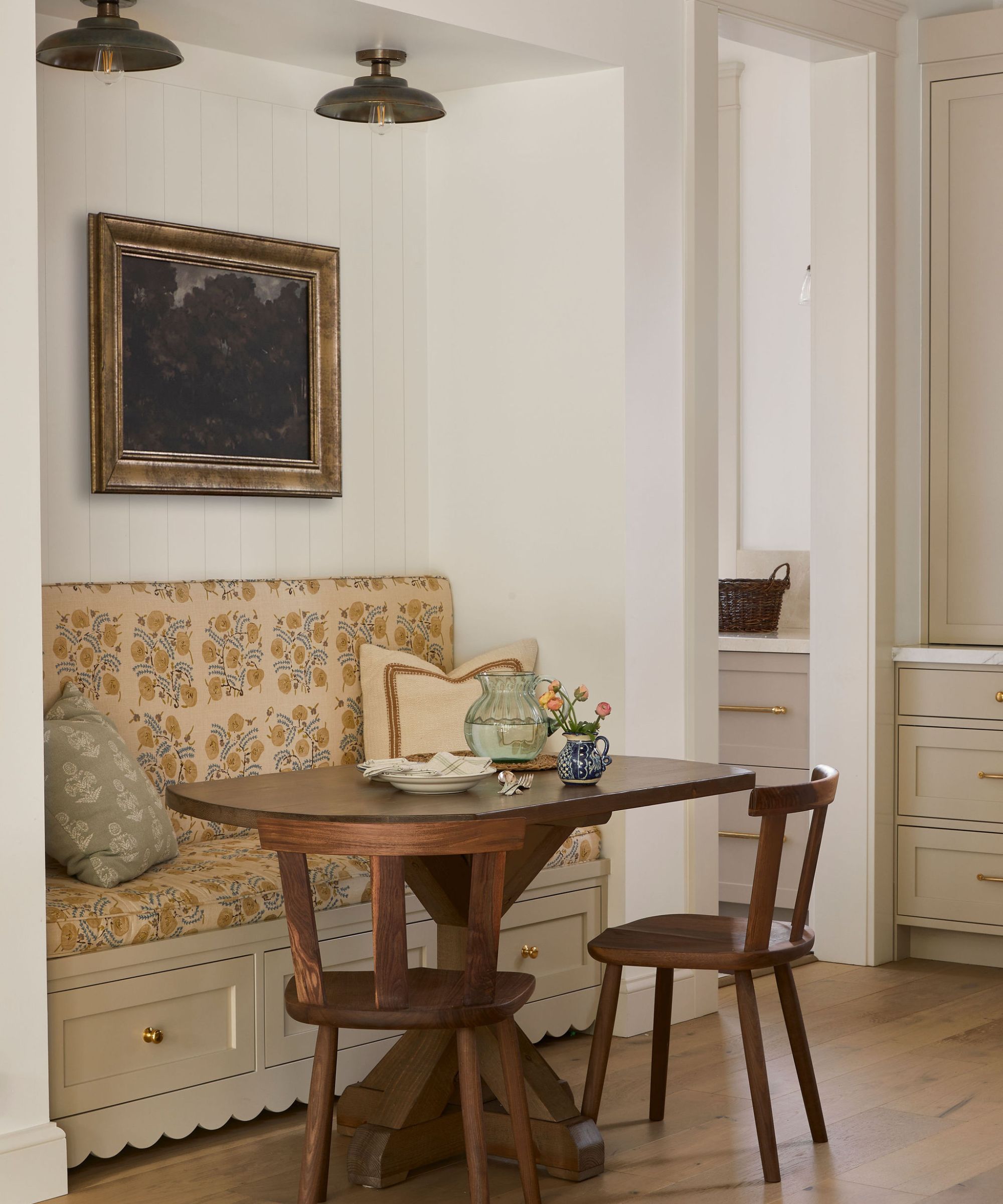 Cozy dining nook with round wooden table and two matching chairs, upholstered banquette seating wih floral fabirc, artwork hung above.