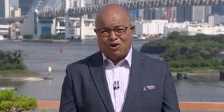 Mike Tirico with a suit on and a purple shirt broadcasting outside in Tokyo at the Olympics.