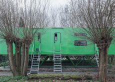 a horizontal green structure sits on diagonal green stilts by Jan Benthem - one of our round up of Dutch houses. there are doors to the structure with metal stairs leading to them. bare trees in front of and behind the structure.