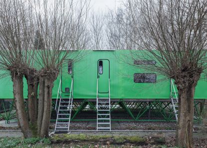 a horizontal green structure sits on diagonal green stilts by Jan Benthem - one of our round up of Dutch houses. there are doors to the structure with metal stairs leading to them. bare trees in front of and behind the structure.