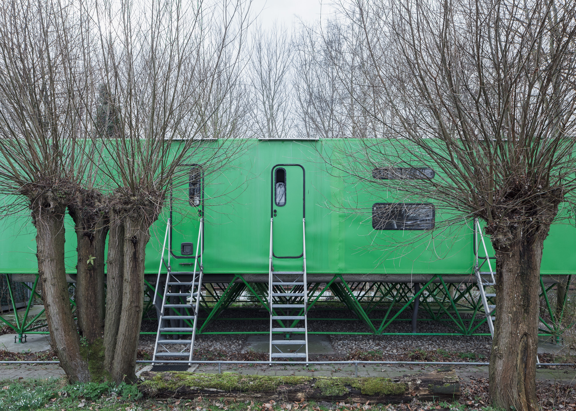a horizontal green structure sits on diagonal green stilts by Jan Benthem . there are doors to the structure with metal stairs leading to them. bare trees in front of and behind the structure.