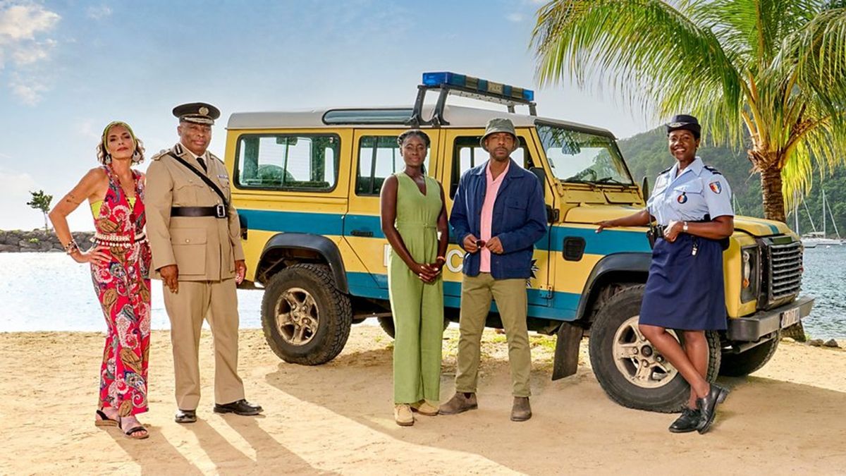 Saint Marie mayor Catherine Bordey (Élizabeth Bourgine) stands beside Commissioner Selwyn Patterson (Don Warrington) and his reporting officers (including, second from right, incoming DI Mervin Wilson) against a tropical Caribbean backdrop in Death in Paradise Season 14.