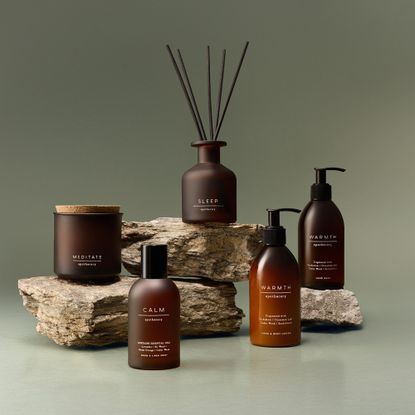 A candle, reed diffuser, room spray and ath body products from the M&S Apothecary range displayed on stacked rocks