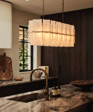 A statement chandelier with rectangular textured glass panels hangs from a chain above a kitchen island. The chandelier emits a warm, golden glow. The island has a dark, marbled stone countertop with a deep brass sink and matching faucet. Behind, dark wooden cabinets and a large window with black metal framing add depth.