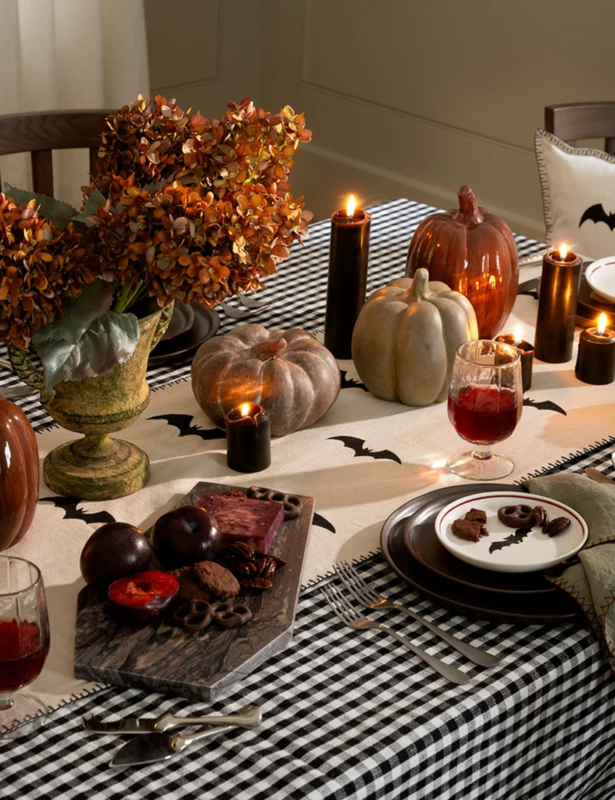 Timeless, richly layered Halloween tablespace featuring a black-and-white gingham tablecloth, marble coffin-shaped cheese board, stacks of chocolate brown plates. and black taper candles aglow.