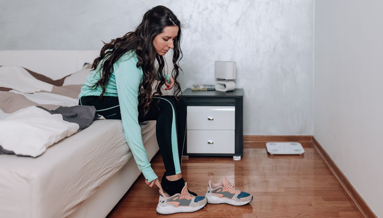 Woman sits on the end of a bed and slips her right foot into a running shoe. She has long black hair and is wearing a running jacket and tracksuit bottoms.