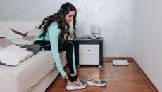 Woman sits on the end of a bed and slips her right foot into a running shoe. She has long black hair and is wearing a running jacket and tracksuit bottoms.