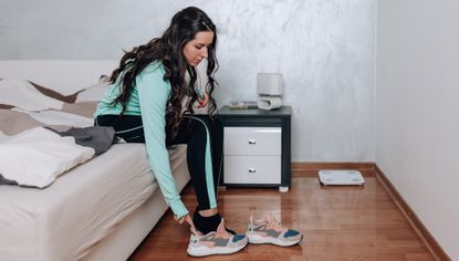 Woman sits on the end of a bed and slips her right foot into a running shoe. She has long black hair and is wearing a running jacket and tracksuit bottoms.