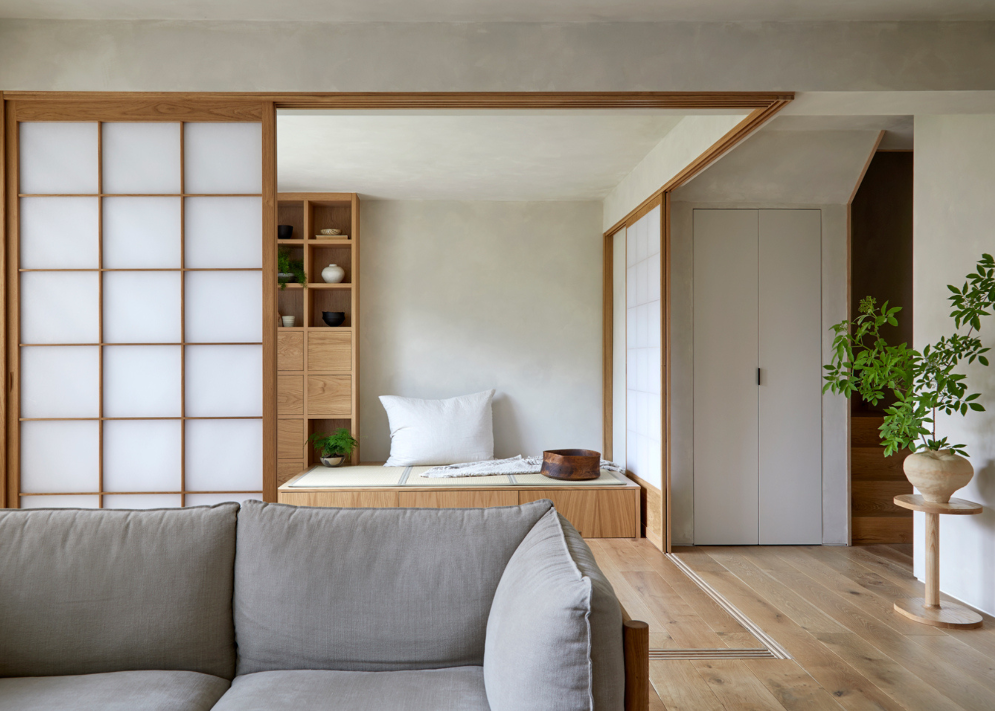 A Japanese living room with a vase of stems on a side table, a gray couch, a sliding rice paper screen, and a bench with a pillow and a bowl