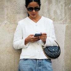 woman wearing white tweed jacket, sunglasses, and jeans looking at her phone and carrying a chanel bag