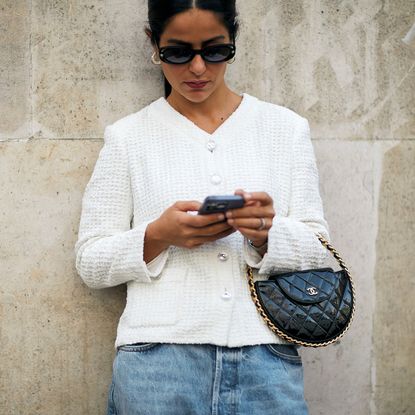 woman wearing white tweed jacket, sunglasses, and jeans looking at her phone and carrying a chanel bag
