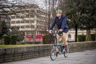 A man in brown trousers and a navy jacket cycles to work on a Brompton folding bike