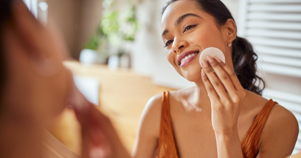 Best toners: Portrait of young woman looking at mirror while cleaning her face with cotton disc in bathroom.