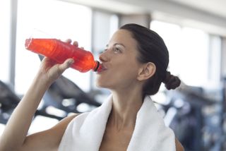 A woman at the gym with a towel around her neck, drinking a red sports drink to re-hydrate after a workout.