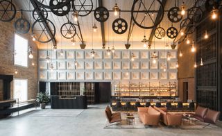 Interior view of The Warehouse Hotel, Singapore featuring grey flooring, oval tables, brown sofas, black chairs at the bar, shelving behind the bar filled with bottles of drinks, a black counter, pendant lights, exposed brick and brown walls and a wall that has a repeating square design. There are also black wheel-like objects suspended from the ceiling