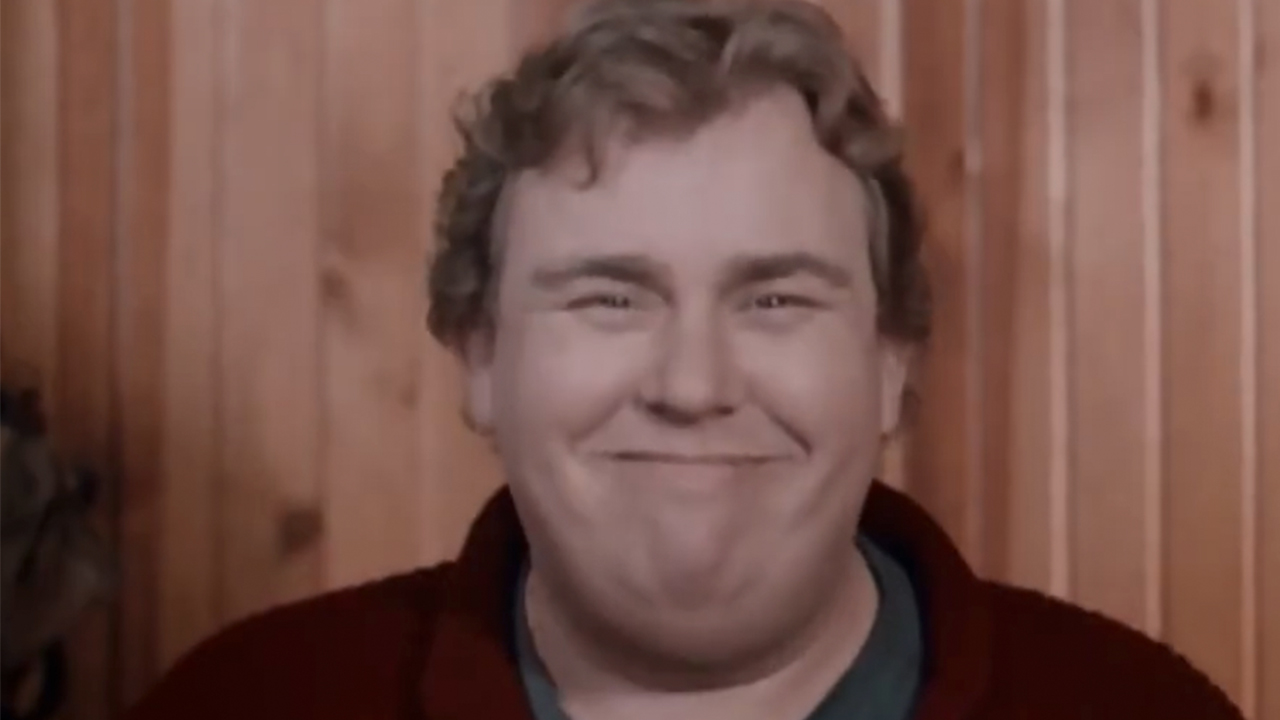 A close up of John Candy smiling in front of a wood wall.