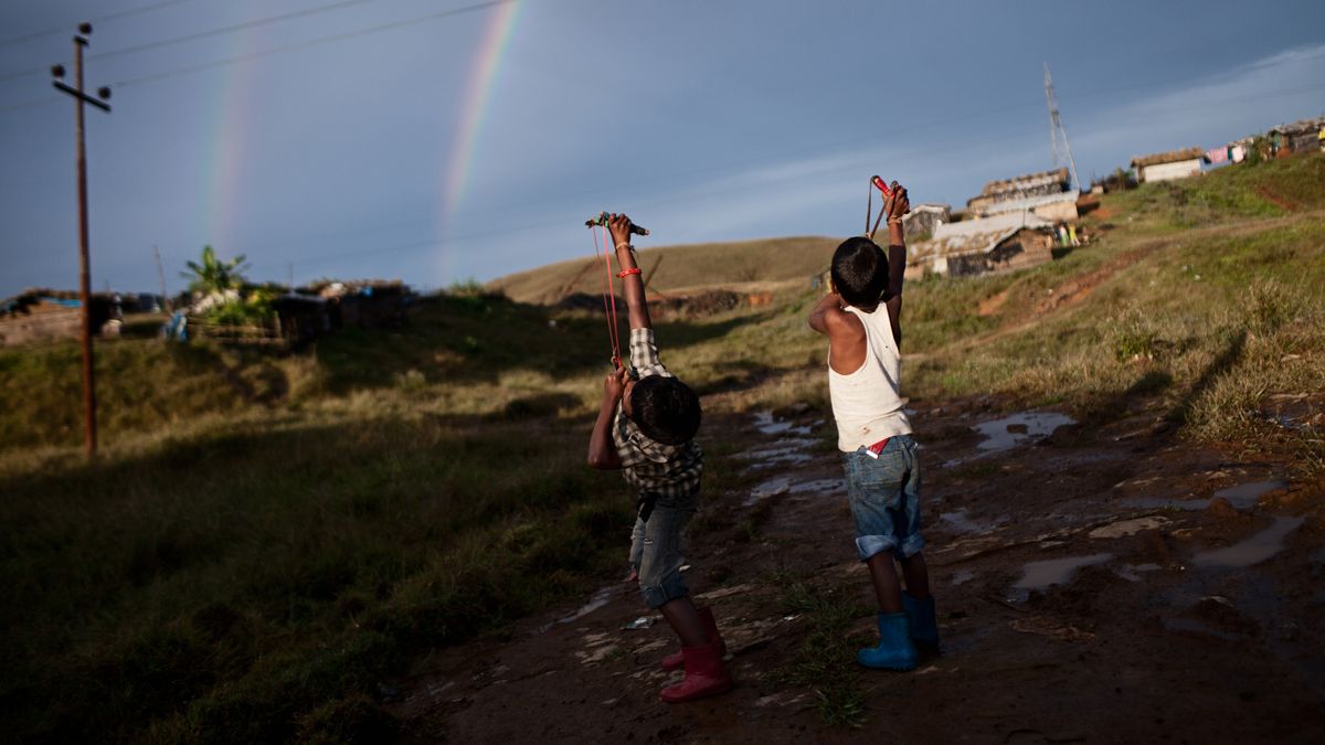  Two boys direct their slingshots to a sky filled with a double rainbow and telephone cables.