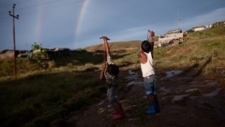  Two boys direct their slingshots to a sky filled with a double rainbow and telephone cables.