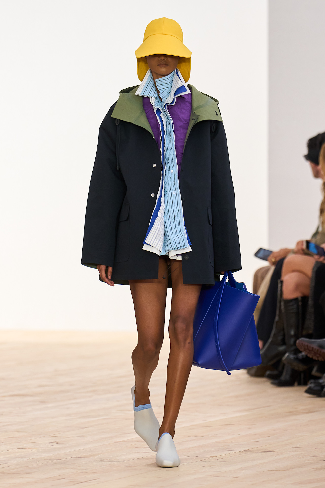 A model at the SS26 Loewe show in Paris wearing a yellow hat, a sporty black jacket, a button-down shirt, and loafers.