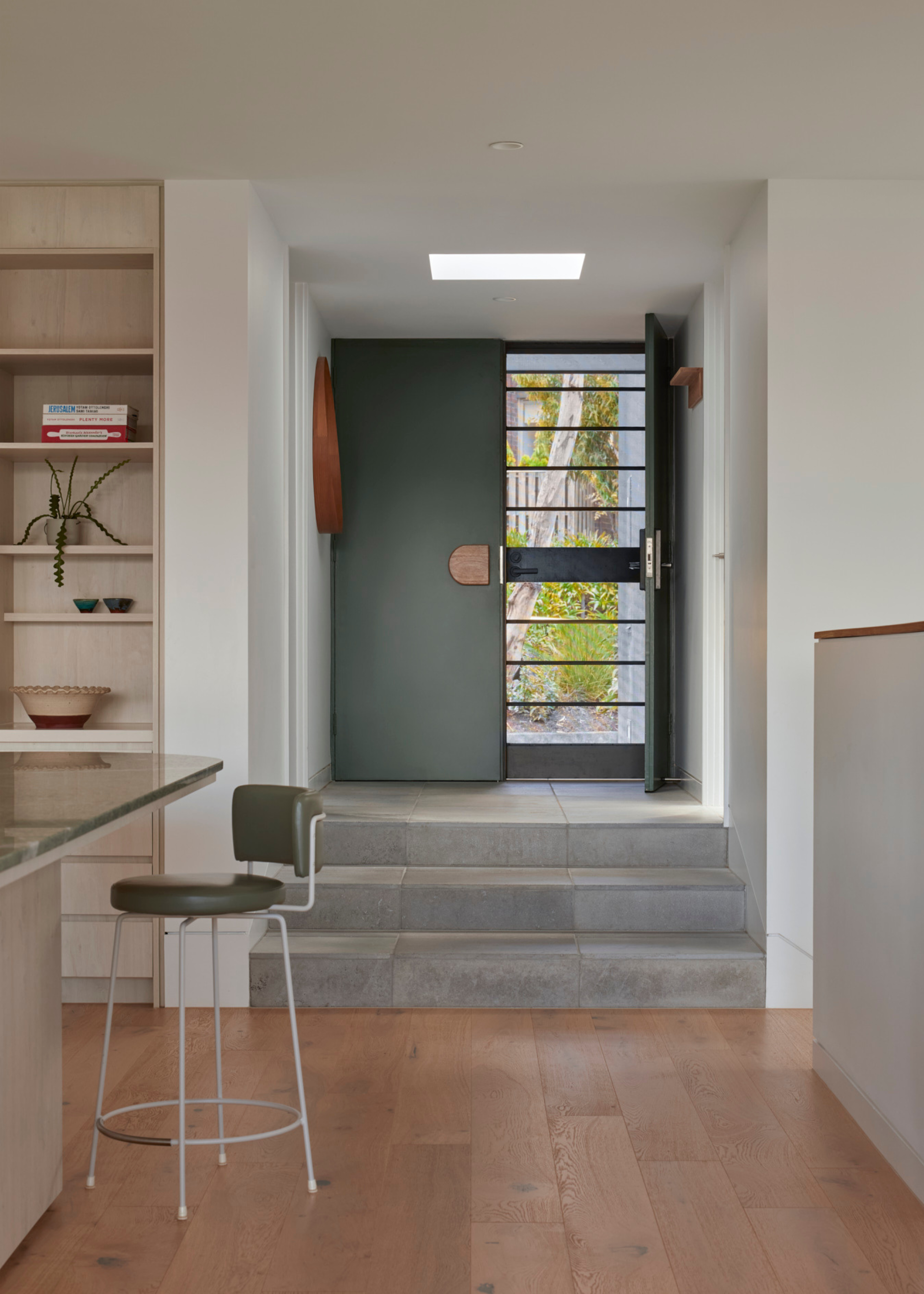 An entryway with a green door, a wood mirror, hooks on the wall, stone steps leading to an open dining space with wood flooring, and a green stool
