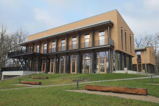 A new office building with two floors predominantly yellow in colour and floor to upper level windows. A large balcony runs along the front of the building. In the foreground is a grass lawn area with wood log benches.