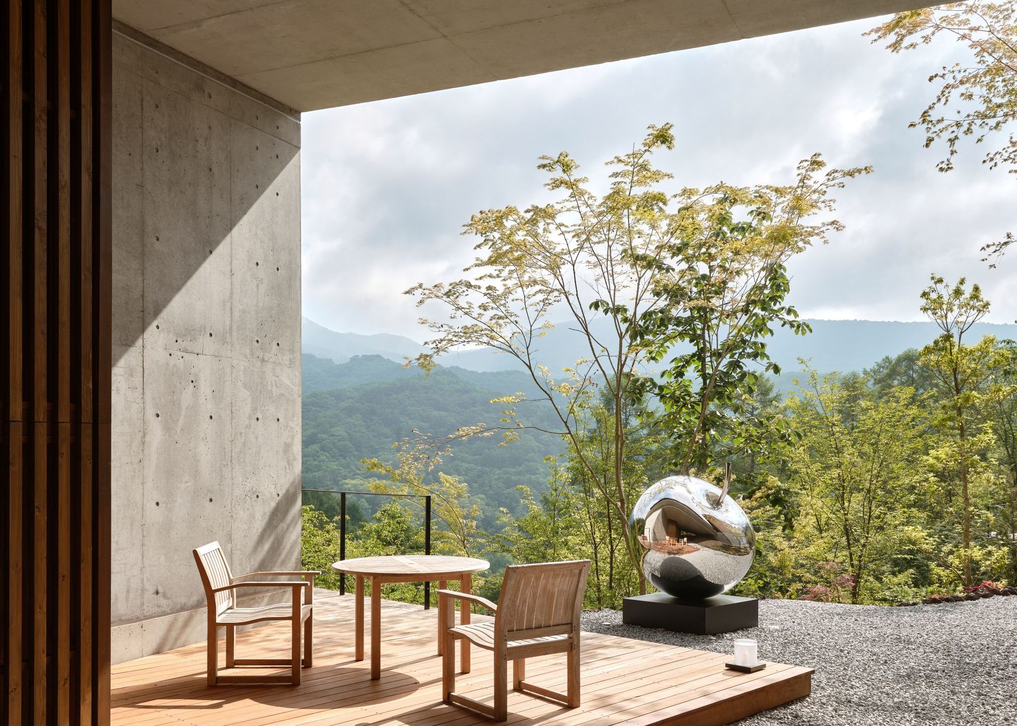 An outdoor seating area in a Japanese home on a slightly raised wooden platform framed by concrete, overlooking the surrounding forest and mountains, with a metallic sculpture in front