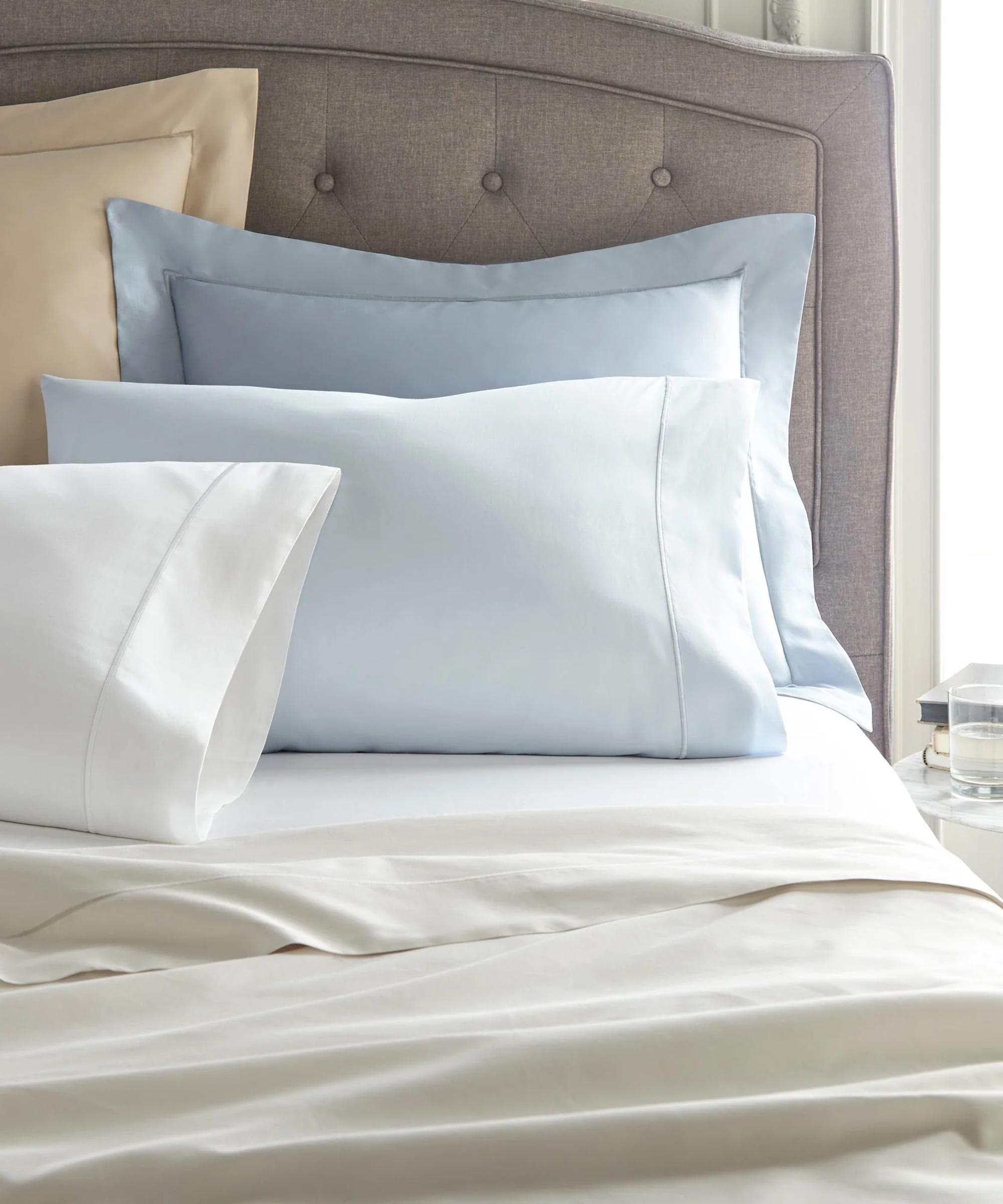 Peacock Alley sateen sheets in various shades of blue and cream styled atop of a bed with a tufted headboard