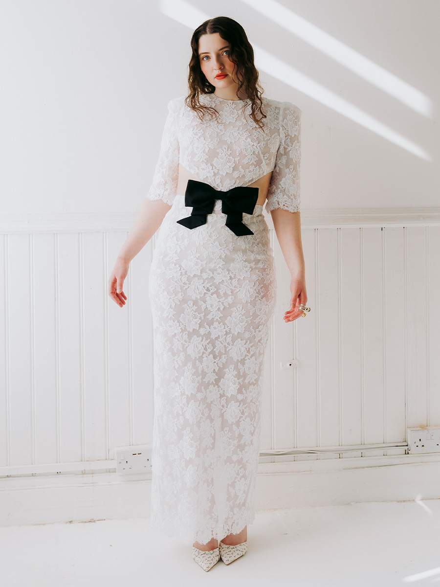 Lucy Halliday photographed by Phill Taylor. She is standing and wearing a white lace dress with center cutout and black bow.