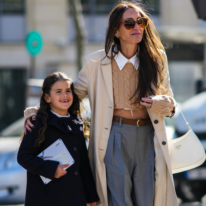 Chloe Harrouche wears sunglasses, a white ripped shirt from Miu Miu, a brown braided wool pullover from Miu Miu, a beige long coat, a brown shiny leather belt from Miu Miu, gray large wide legs pants, white leather ripped sneakers, and is seen with her daughter, outside Miu Miu , during Paris Fashion Week - Womenswear F/W 2022-2023, on March 08, 2022 in Paris, France.