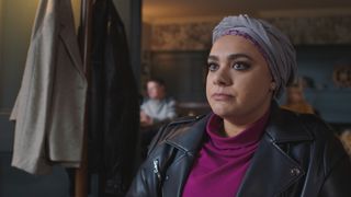 Sarah Seggari as nurse Rida Amaan in a pub looking stern and wearing a lilac headscarf, purple polo neck and black jacket.