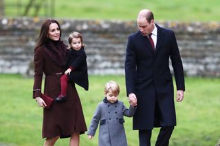 Princess Kate wearing a brown coat with a fur collar holding Princess Charlotte, walking outside next to Prince William who is holding hands with Prince George