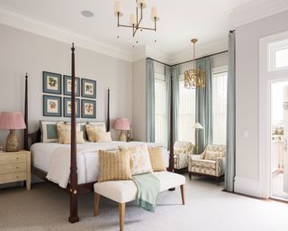 A gallery bedroom accent wall with square prints above a four poster bed in a traditional, neutral bedroom scheme with pale blue drapes and creamy yellow and pale pink accents.