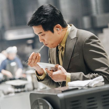 A Korean man (chef Ahn Sung-jae) tastes a meal with chopsticks. Chefs work at cook stations the blurred background, in a still from Netflix's 'Culinary Class Wars.'